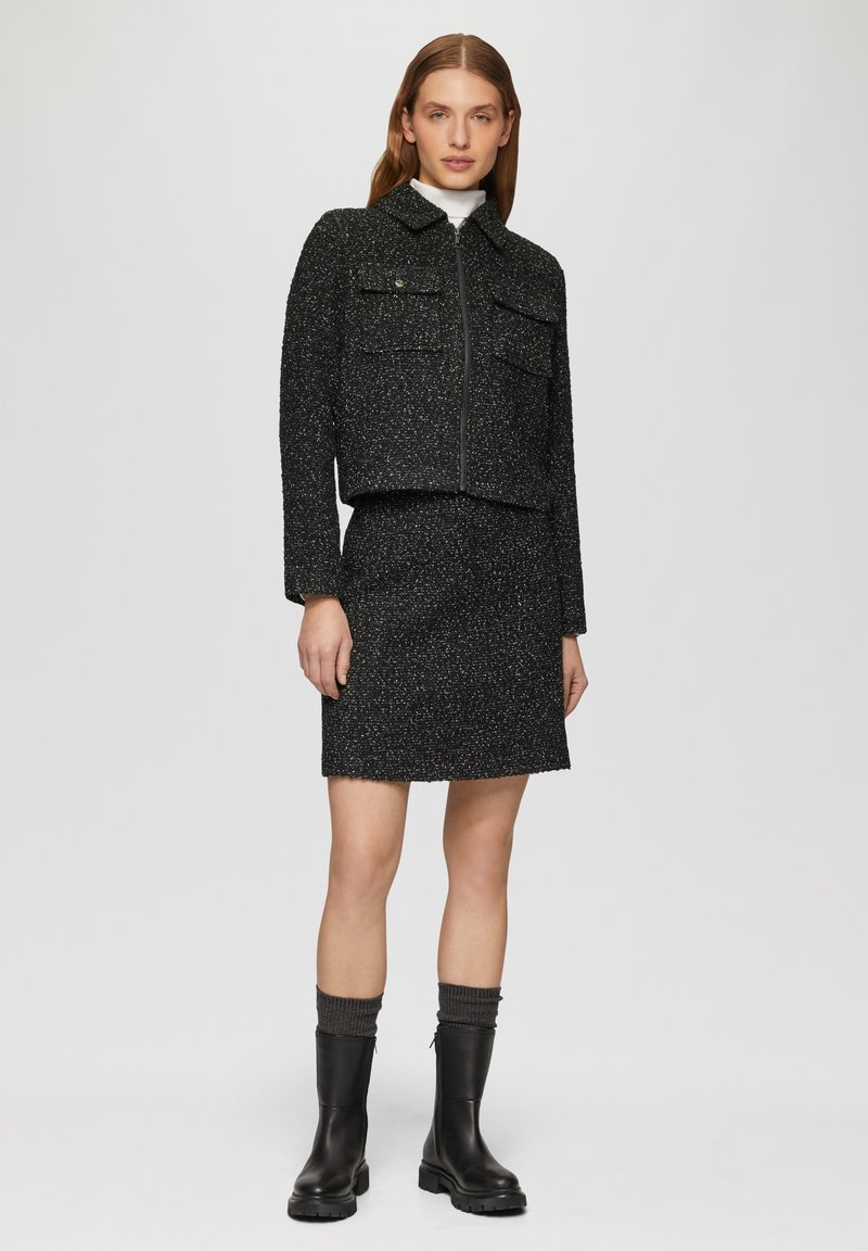 Schwarze Tweed-Cropped-Jacke mit zwei Fronttaschen, kombiniert mit einem passenden Rock. Das Model trägt knielange schwarze Stiefel mit grauen Abschlüssen.