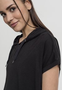 Ung kvinna med långt brunt hår i en svart kortärmad hoodie som ler, med en tunn fläta hängande vid hennes ansikte, mot en vit bakgrund.
