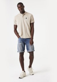 Beige poloshirt met een klein logo, gecombineerd met lichtblauwe denim shorts met rafelige randen. Witte sneakers maken de outfit compleet.