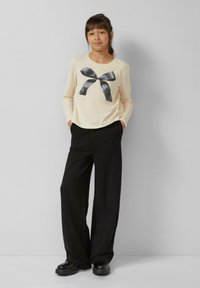 Crèmekleurig shirt met lange mouwen en een zwart polkadotstrikje als afbeelding, gecombineerd met zwarte, hooggesneden broek met wijd uitlopende pijpen en stoere zwarte schoenen.