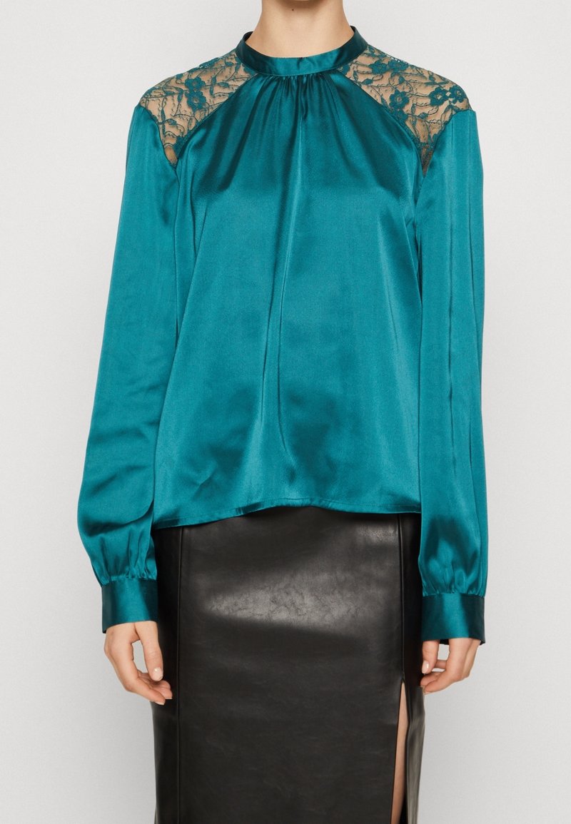 Blouse en satin bleu sarcelle avec panneaux en dentelle sur les épaules, encolure froncée et manches longues. Associée à une jupe en cuir noire avec une fente.