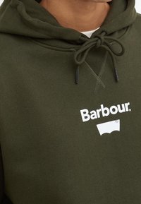 Gros plan d'une personne portant un pull à capuche vert olive avec des cordons noués et le logo Barbour blanc sur la poitrine.