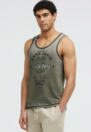 Mann mit lockigem dunklem Haar, der ein grünes Camp David Tanktop und beige Hose mit Kordelzug trägt, steht mit einer Hand in der Tasche.