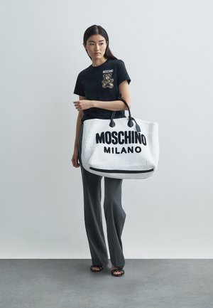Femeie îmbrăcată cu un tricou negru cu urs de pluș și pantaloni gri, ținând o geantă mare albă MOSCHINO MILANO cu mânere negre.