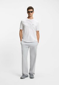 White short-sleeve shirt with gray graphic design and logo, paired with light gray trousers and gray athletic shoes, finished with dark sunglasses.