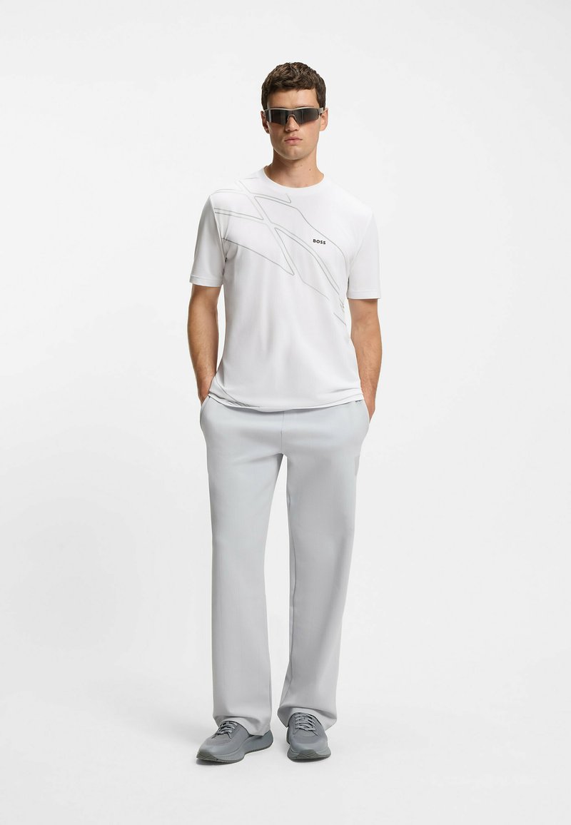 White short-sleeve shirt with gray graphic design and logo, paired with light gray trousers and gray athletic shoes, finished with dark sunglasses.