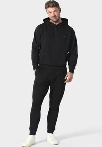 Felpa nera con cappuccio e tasca a marsupio, abbinata a pantaloni della tuta affusolati coordinati. Realizzata in tessuto morbido, mostrata con sneakers bianche.