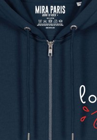 Tums zilā džemperis ar rāvējslēdzēju, izgatavots no 100% kokvilnas, ar metāla rāvējslēdzēju, auklām un grafisku tekstu "Born to Rock" baltā un sarkanā krāsā.