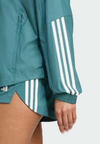 Teal windbreaker with white triple stripes on the sleeves, elastic cuffs, and a side pocket detail. Pairing with a coordinating short.