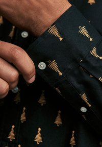 Chemise noire à boutons ornée de motifs d'arbres dorés, avec des détails de poignets et de boutons visibles. Le tissu semble texturé et durable.