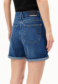 Shorts en denim bleu foncé avec un ourlet roulé, présentant un design de poche arrière et une étiquette noire au niveau de la taille. Texture en coton.