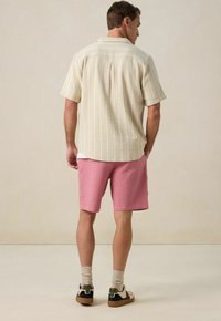 Heren kortarmig shirt in roomkleur met verticale strepen, gecombineerd met textuur roze shorts. Sokken en casual schoenen maken de outfit compleet.