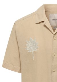 Camisa de manga corta beige en tejido ligero con cuello, que presenta un diseño bordado de una hoja verde claro en el lado izquierdo del pecho.