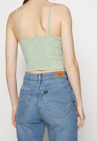Mujer que lleva un top corto de canalé verde claro con tirantes delgados y unos jeans azul claro de tiro alto de Lee, mostrada de espaldas contra un fondo liso.