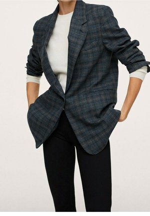 Personne portant un blazer à carreaux foncé avec les manches retroussées sur une chemise blanche à manches longues, associée à un pantalon noir, mains dans les poches.