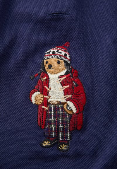 Broderi av en bjørn som bærer en rød frakk, mønstrete hatt og rutete bukser på en marineblå bakgrunn. Har detaljerte sting og tekstur.