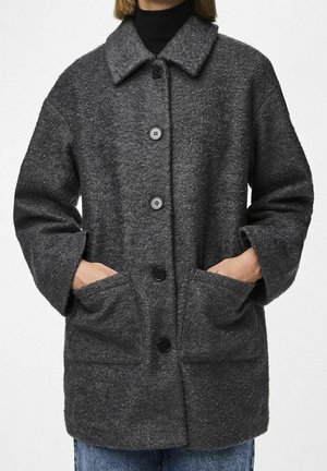 Personne portant un manteau en laine gris foncé boutonné avec de grandes poches avant et un large col, mains placées à l'intérieur des poches.