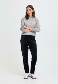 Grå strikket genser med høy krage, kombinert med svarte kordfløyelsbukser med innsnevring. Modellen har på seg hvite joggesko med svarte detaljer.