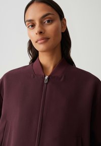 Giacca bomber borgogna con colletto rialzato, dotata di zipper frontale e tasche laterali; texture liscia e linee pulite.