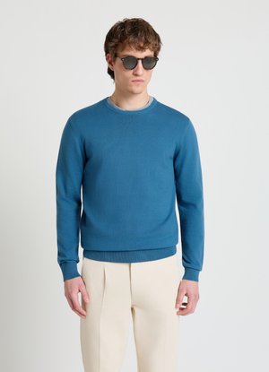 Jeune homme portant des lunettes de soleil noires, un pull col rond bleu sarcelle et un pantalon plissé beige, debout devant un fond clair uni.