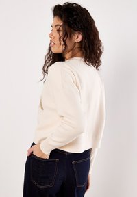 Pull crème, court, avec un col arrondi et des manches longues, featuring un détail texturé. Associé à un jean en denim foncé.