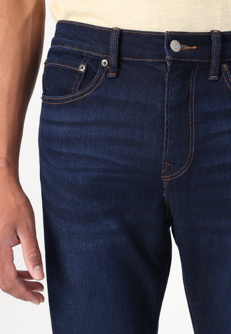 Jeans de mezclilla azul oscuro con un corte slim. Presenta costuras naranjas, bolsillos frontales y un cierre de botón de metal en la cintura.