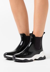 Bottines noires brillantes avec panneaux élastiques sur les côtés, semelles en caoutchouc blanches et semelles texturées noires pour une meilleure adhérence.