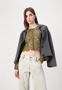 Top court vert olive en dentelle avec motif floral et fermeture à boutons, superposé sous un blazer en laine grise. Jean large en denim clair.