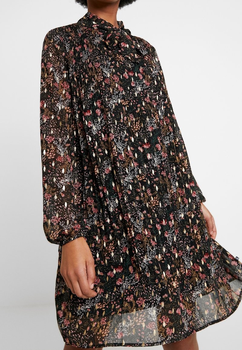 Femme portant une robe noire à manches longues avec un imprimé floral rose et beige et un nœud au cou, tenant l'ourlet de la robe.