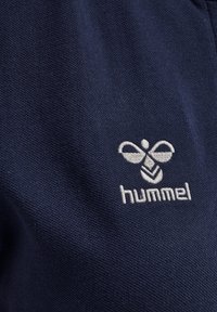 Marineblåt stof med en struktureret vævning, der har et hvidt broderet "hummel"-logo og bi-ikon, placeret på venstre side.