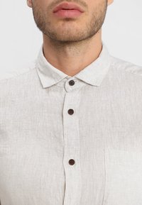 Camisa de lino en beige claro con botones, que presenta un cuello puntiagudo, botones marrón oscuro y un bolsillo en el pecho. Textura suave y diseño casual.