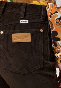 Bruna kordfloddsbyxor med en läderetikett med "Wrangler" präglat. Synlig sömnadsdetaljer och metallnitar på fickorna.