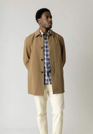 Homme portant un manteau boutonné beige sur une chemise à carreaux bleue et blanche avec un pantalon écru, debout devant un fond clair uni.