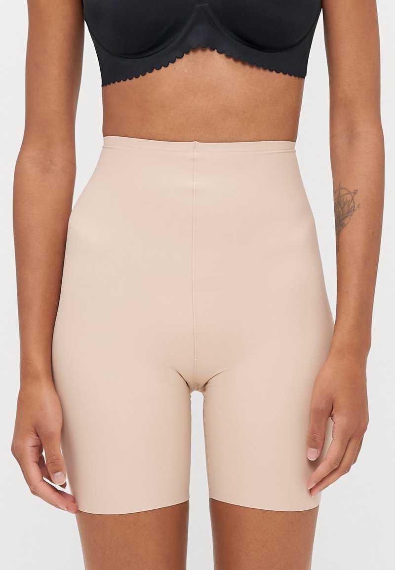 Naine kannab kõrge vöökohtaga beeže vormivaid lühikesi pükse ja musta rinnahoidjat, tema vasakul eelkäel on nähtav tätoveering.