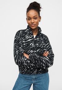 Μαύρο και άσπρο φλις πουλόβερ με ρίγες ζέβρας, ψηλό γιακά και κλείσιμο με μισό φερμουάρ· συνδυασμένο με μπλε τζιν παντελόνι. Μαλακή υφή.