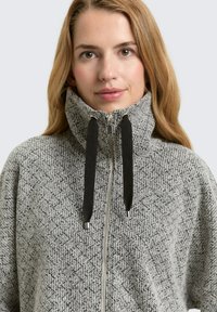 Vrouw in een grijze fleece jas met hoge kraag, voorzien van een rits en twee zwarte trekkoorden, die naar voren kijkt tegen een effen achtergrond.