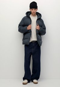 Kurtka puchowa w ciemnoszarym kolorze, z kapturem i elastycznymi mankietami, w połączeniu z beżowym swetrem i luźnymi ciemnymi dżinsami.