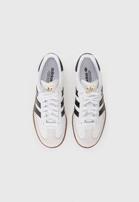 White athletic shoes with three black stripes, padded collar, and brown rubber sole. Features gold logo on the tongue and textured suede accents.