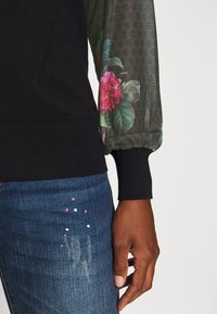 Höger arm i svart tröja med genomskinlig blommig ärm och manschett, vilande bredvid blå jeans med små rosa och vita färgfläckar.