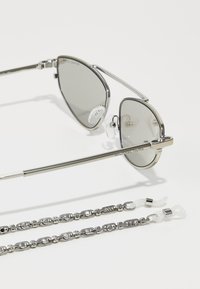 Occhiali da sole in metallo argentato con lenti grigie, caratterizzati da una forma elegante a occhio di gatto e con naselli, accompagnati da una catena argentata con estremità in plastica.