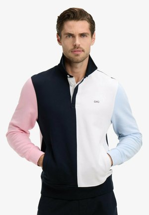 Homme aux cheveux bruns portant un pullover à manches longues avec une fermeture éclair à moitié, manche gauche rose, devant bleu marine et blanc, et manche droite bleu clair.