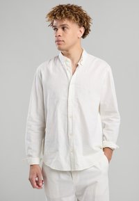 Giovane con capelli ricci che indossa una camicia bianca con i bottoni sbottonata e pantaloni bianchi, che guarda a sinistra con una mano in tasca.