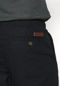 Nahaufnahme von schwarzen Hosen mit einer aufknöpfbaren Gesäßtasche und einem braunen Indicode-Markenpatch, kombiniert mit einem dunkelgrünen, strukturierten Hemd.