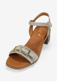 Sandalia de ante beige con un tacón bajo en bloque, que presenta una correa ancha con una hebilla rectangular dorada y una plantilla marrón marcada con la talla 36.