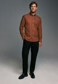 Camisa marrón de botones con cuello, pantalones negros y zapatos negros. Tela suave con botones blancos. Diseño simple, silueta ajustada, fondo neutro.
