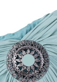 Un tissu turquoise présente de doux plis, accentués par un broche ornementale en argent avec des motifs floraux intricats et un miroir central.