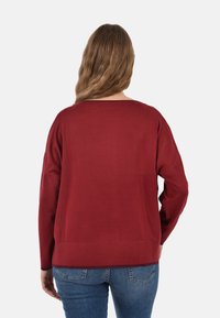 Maglione rosso a maglia con vestibilità ampia, scollatura larga e maniche lunghe. Tessuto liscio con dettagli a coste sull'orlo. Indossato con jeans blu.