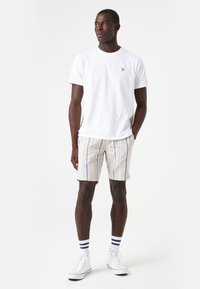Camiseta de algodón blanca con un pequeño logo, combinada con pantalones cortos a rayas beige y zapatillas blancas con detalles en azul marino. El modelo se encuentra de pie contra un fondo claro.