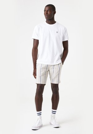 White cotton t-shirt with a small logo, paired with beige striped shorts, and white sneakers with navy accents. Model stands against a light background.
