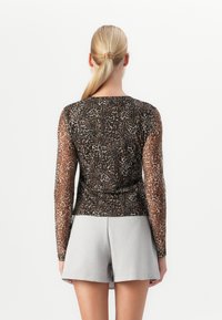 Woman with blonde ponytail wearing a sheer leopard-print long-sleeve top and light gray shorts, shown from the back against a white background.